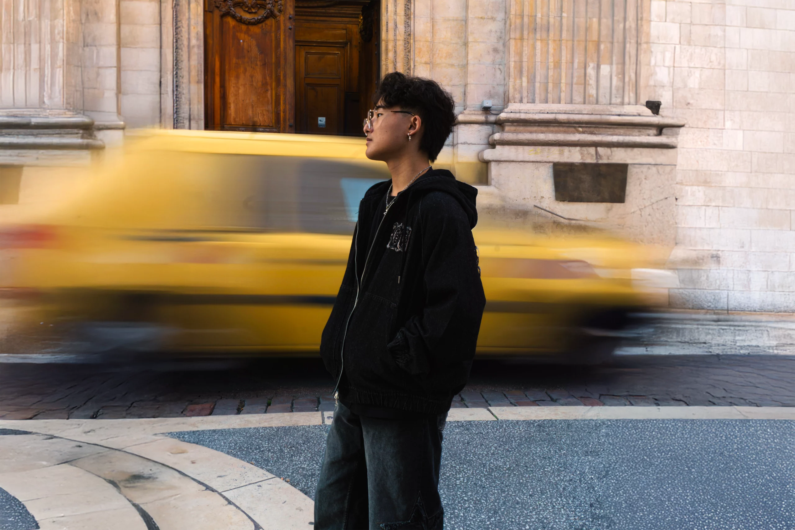 Doomin Park, aka Aeruk, beside a stone façade as a yellow car passes by.